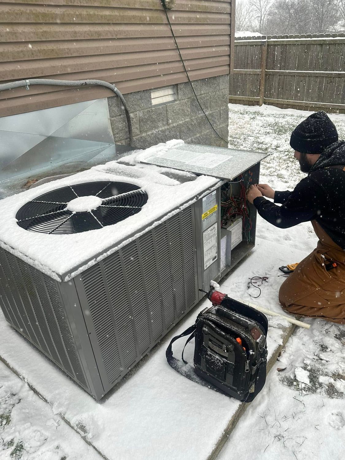 Worker repairing outdoor HVAC unit in snow beside a house