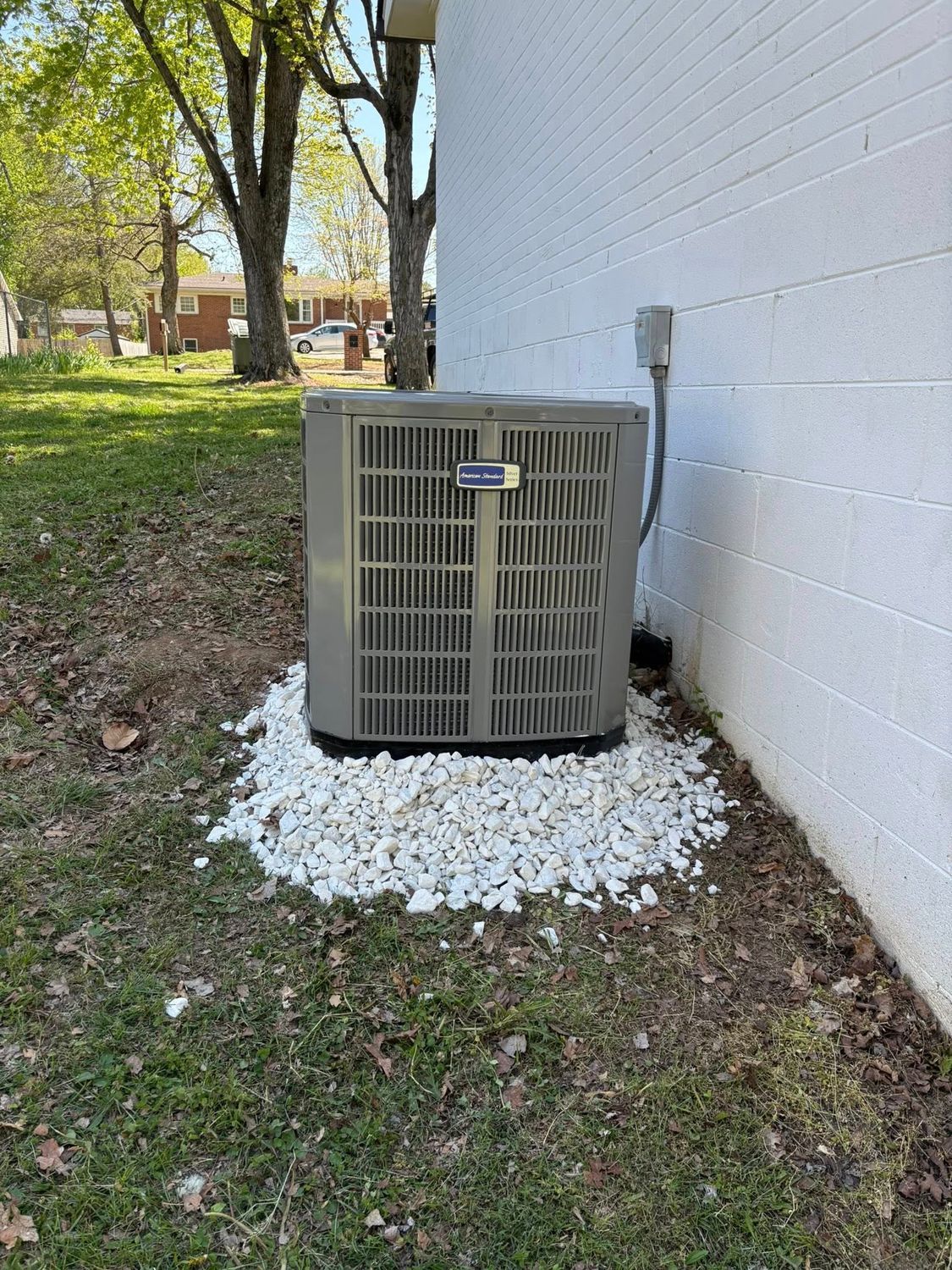 Outdoor air conditioner unit beside a white brick wall, set on white gravel near a grassy yard.