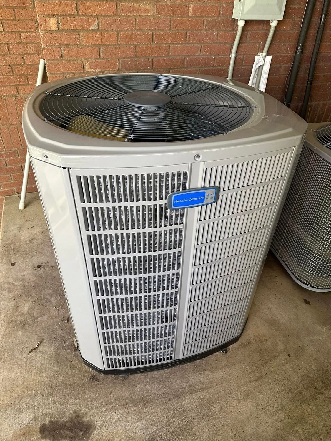 Outdoor air conditioner unit on a concrete patio beside a brick wall, with a fan grille on top.