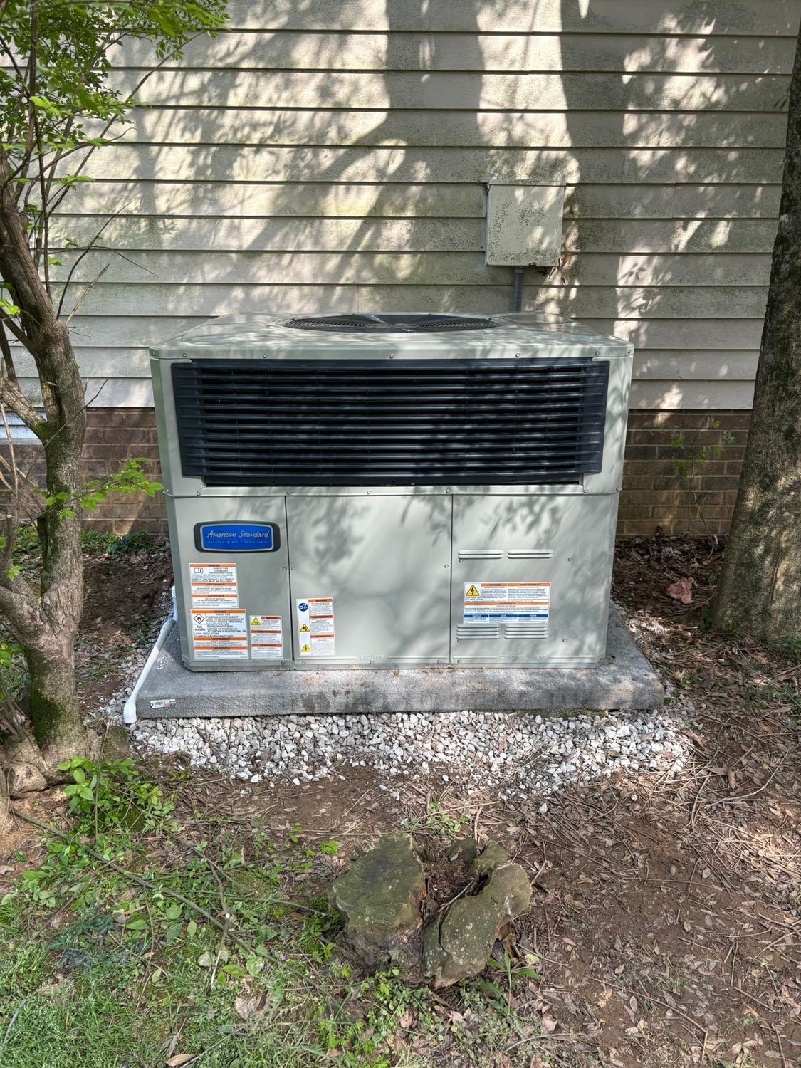Outdoor HVAC unit on a concrete pad beside a house, with bushes and gravel around it.
