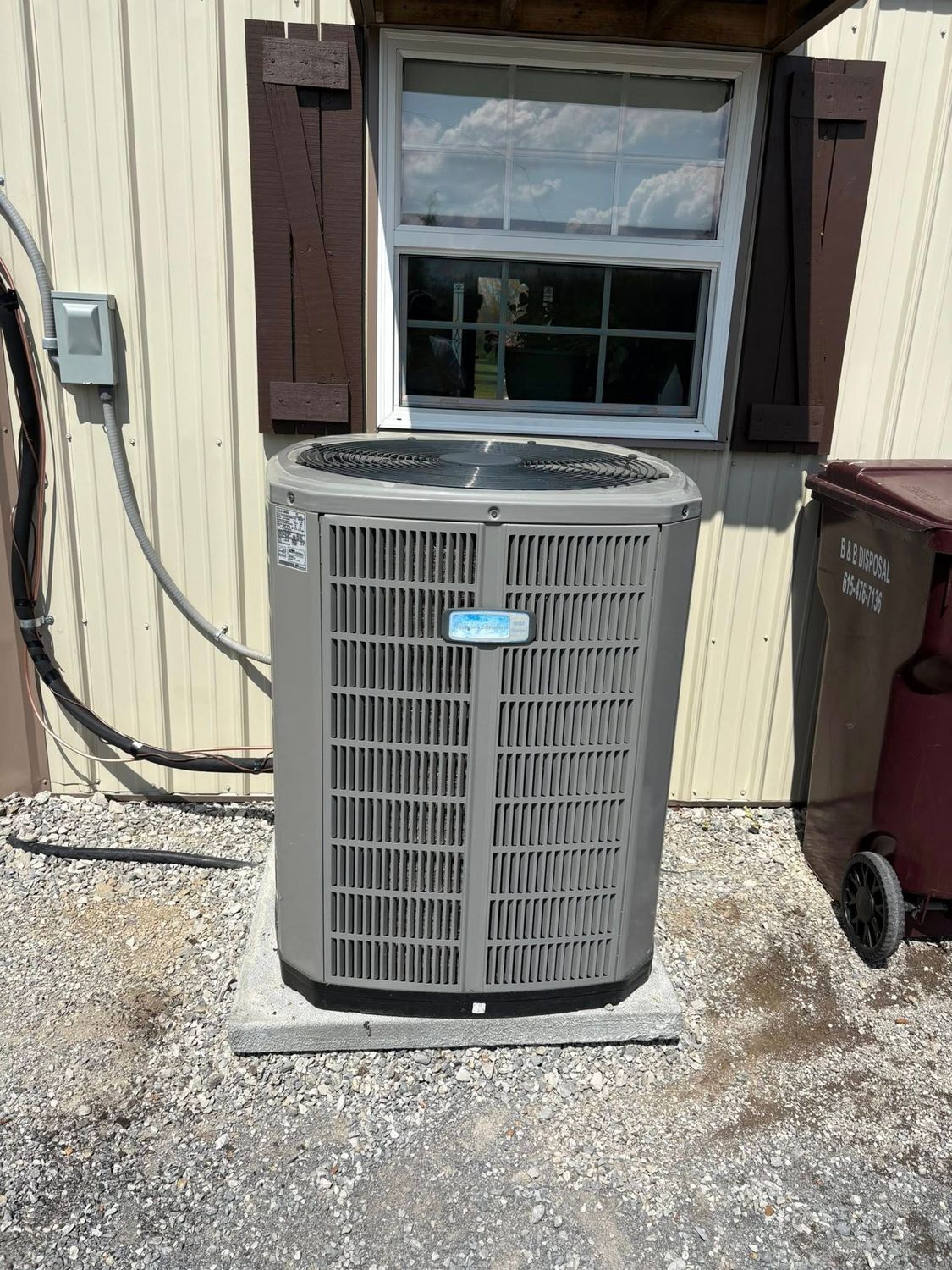 Outdoor central AC unit beside a house window with brown shutters and utility cables on the wall