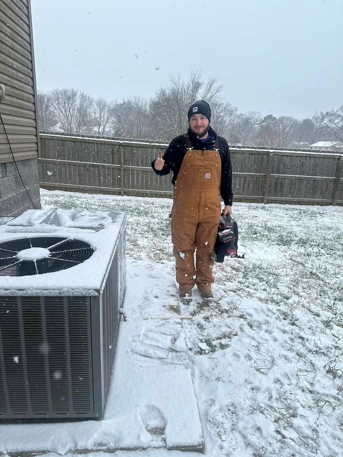 Person in brown overalls standing in a snowy yard, giving a thumbs-up beside an outdoor HVAC unit.