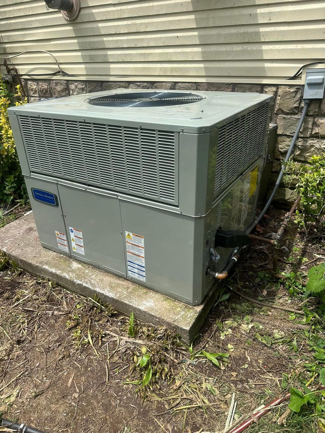 Outdoor HVAC unit beside a house on a concrete pad, with beige siding and surrounding plants and dirt.
