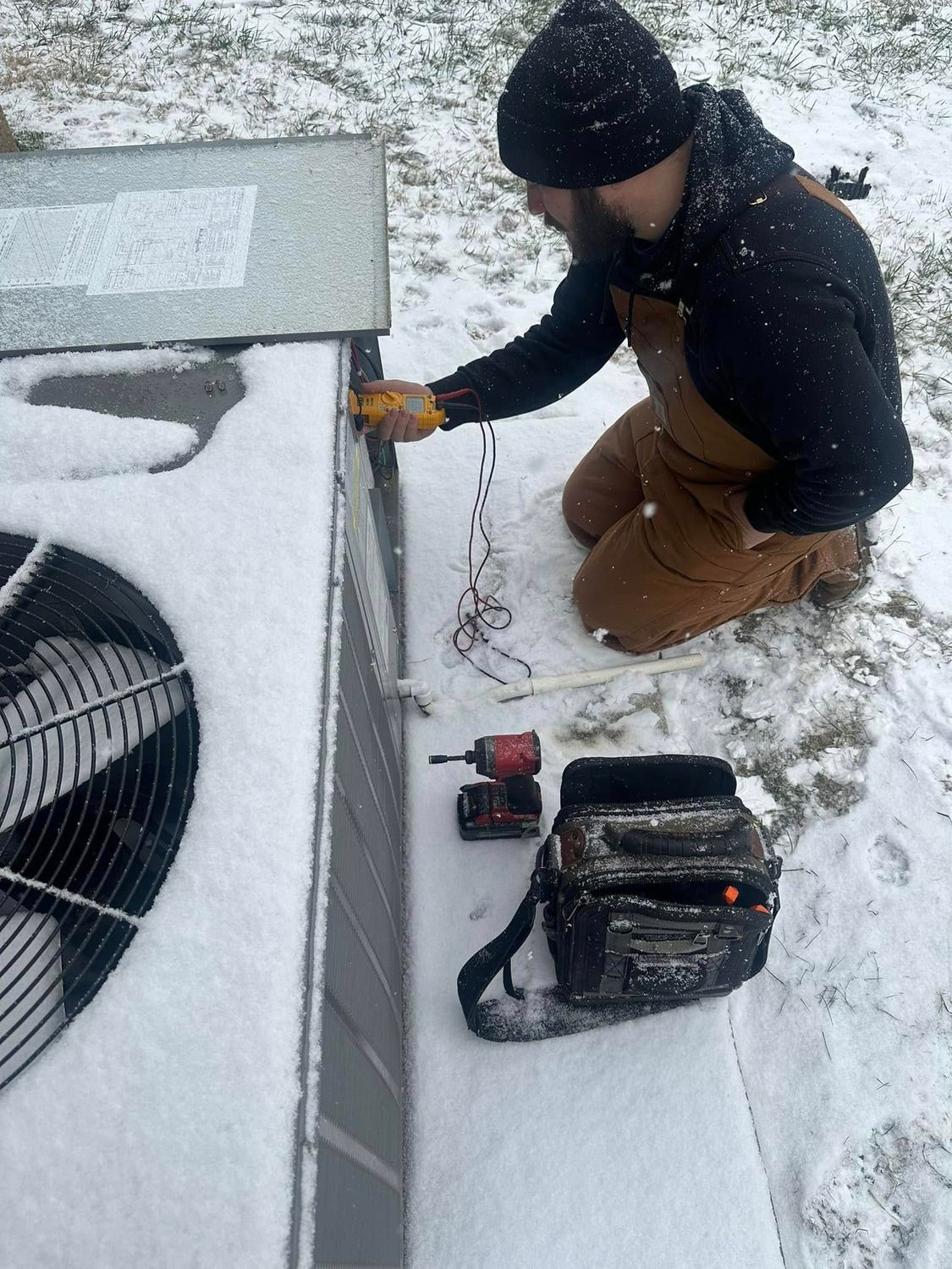 Person kneeling in snow, measuring a rooftop edge with a tape measure near an HVAC unit and tool bag.