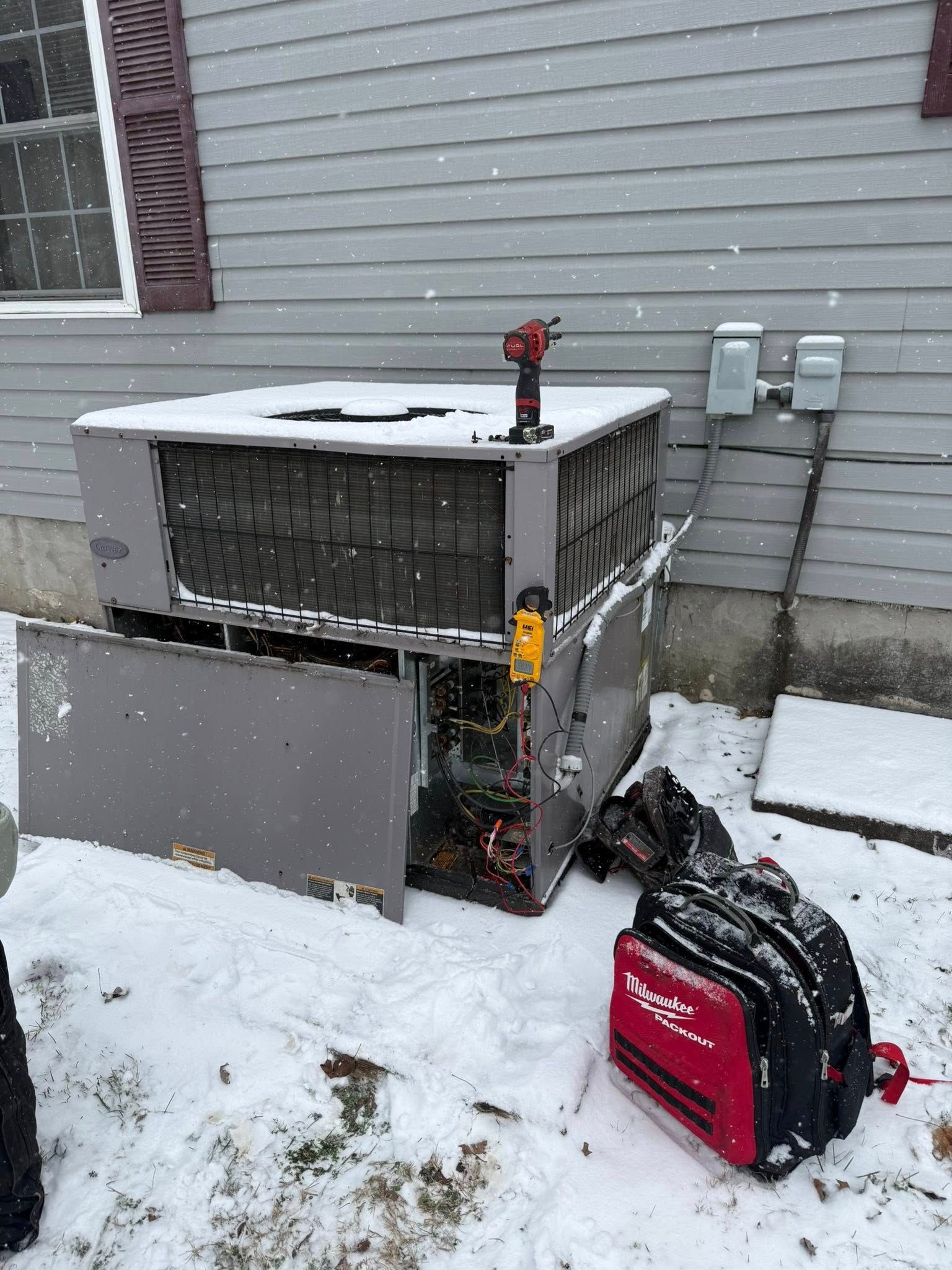 Outdoor air conditioner unit in snow beside a house, with a red tool bag nearby.