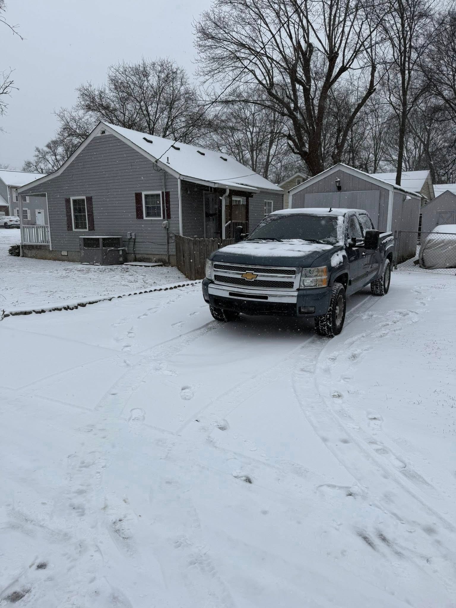 Snowy residential street with a black SUV parked in front of a small house and shed.
