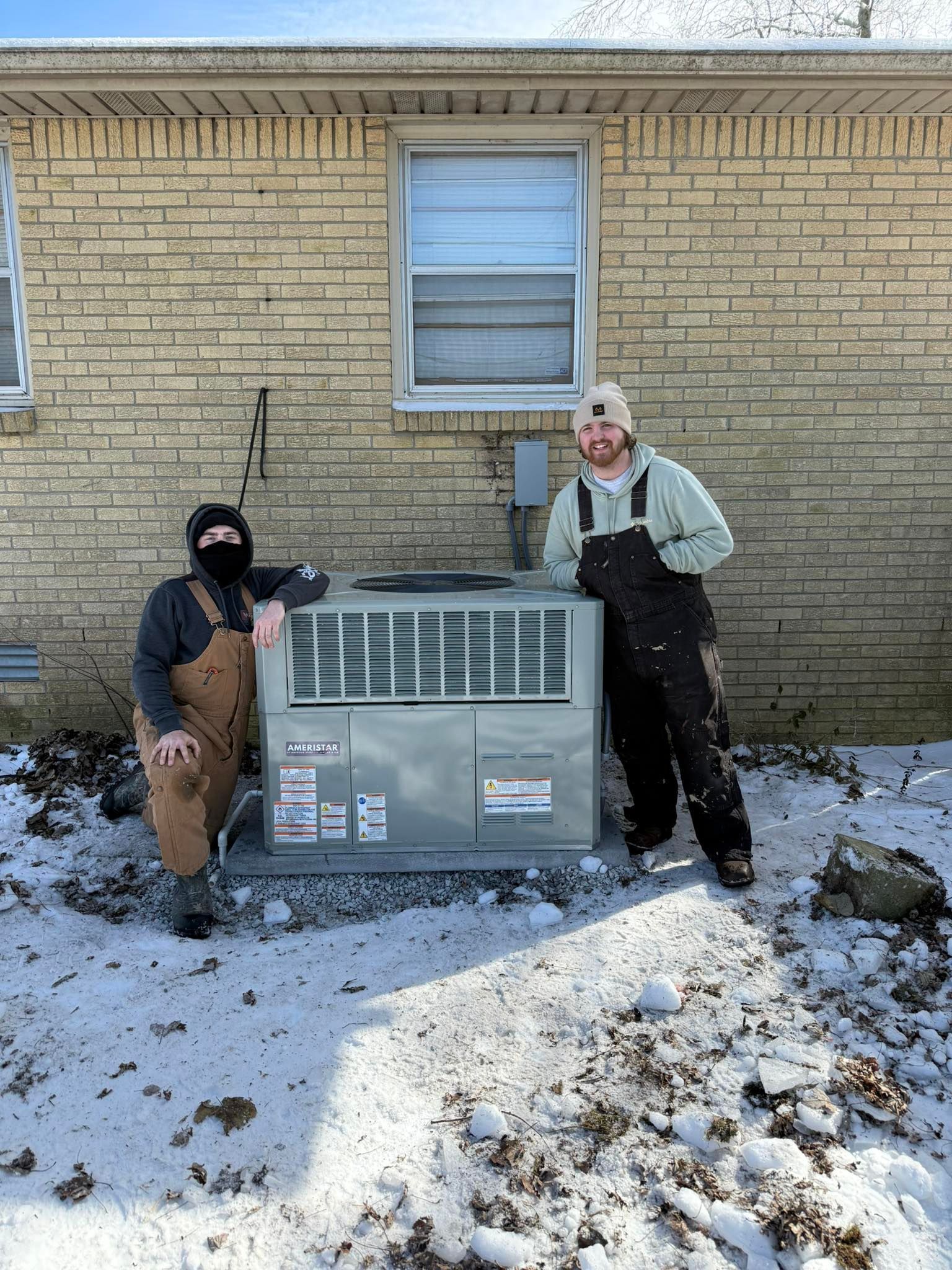 Two workers standing beside a large outdoor HVAC unit against a brick house in snow