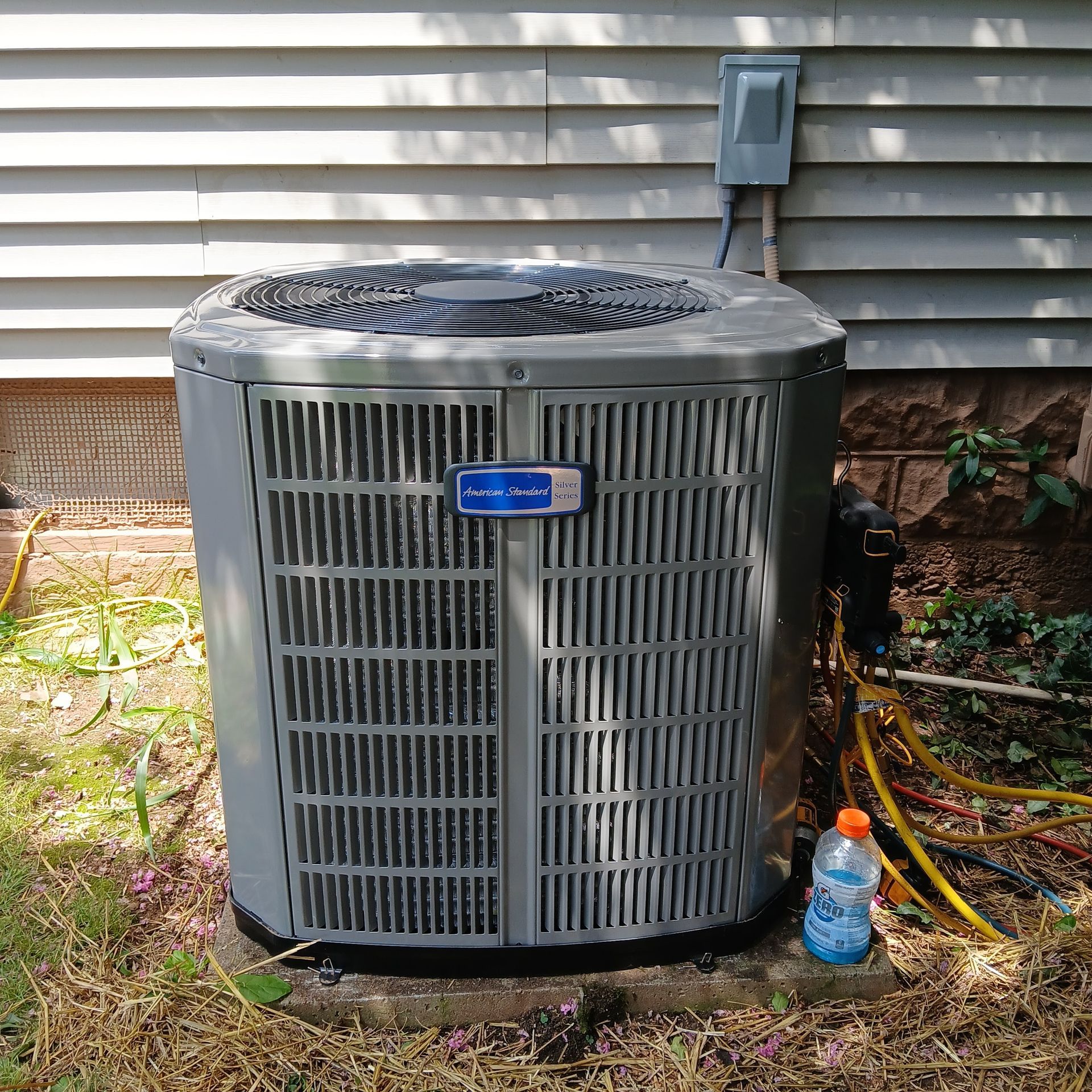 Outdoor air conditioner condenser unit beside house siding with hoses and a water bottle nearby
