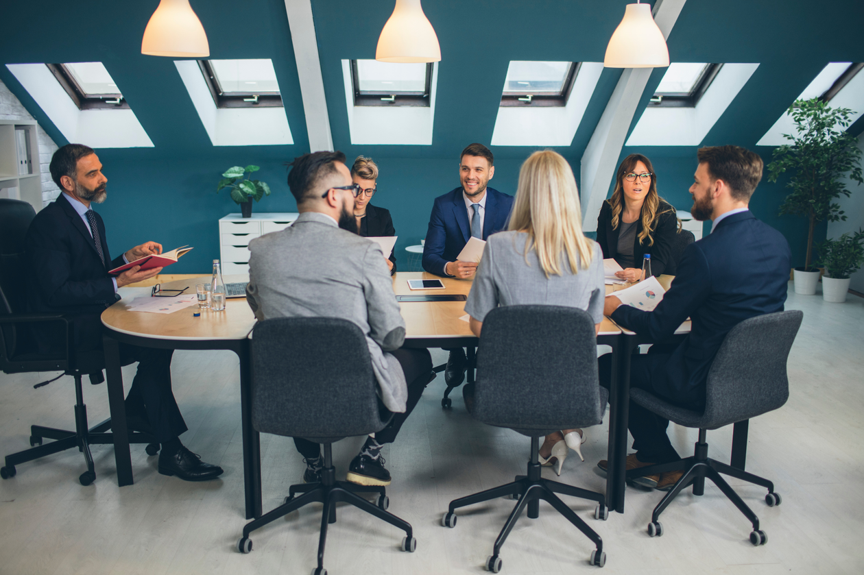 Reunión de negocios alrededor de una mesa de conferencias en una oficina luminosa, con seis personas sentadas conversando.