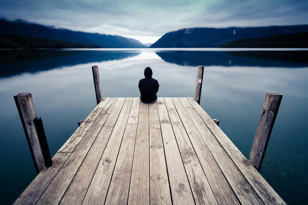 Una persona sentada al final de un muelle de madera contemplando un lago tranquilo y montañas lejanas al atardecer.