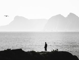 Silueta de una persona en una costa rocosa frente a un mar en calma con montañas brumosas al fondo.
