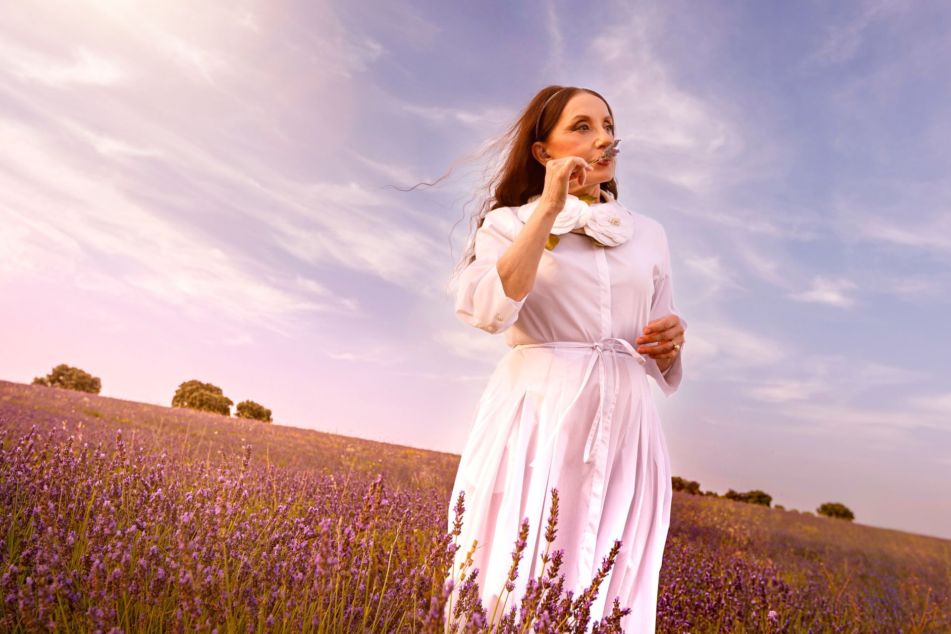 Luz Casal, con vestido blanco, sostiene una flor de lavanda cerca del rostro entre los campos de Brihuega.