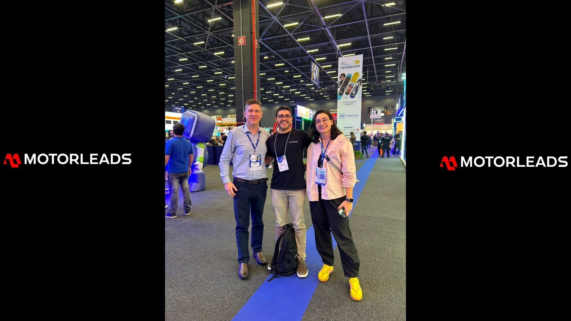 Three people posing at an indoor expo, standing on a blue carpet with bright overhead lights.