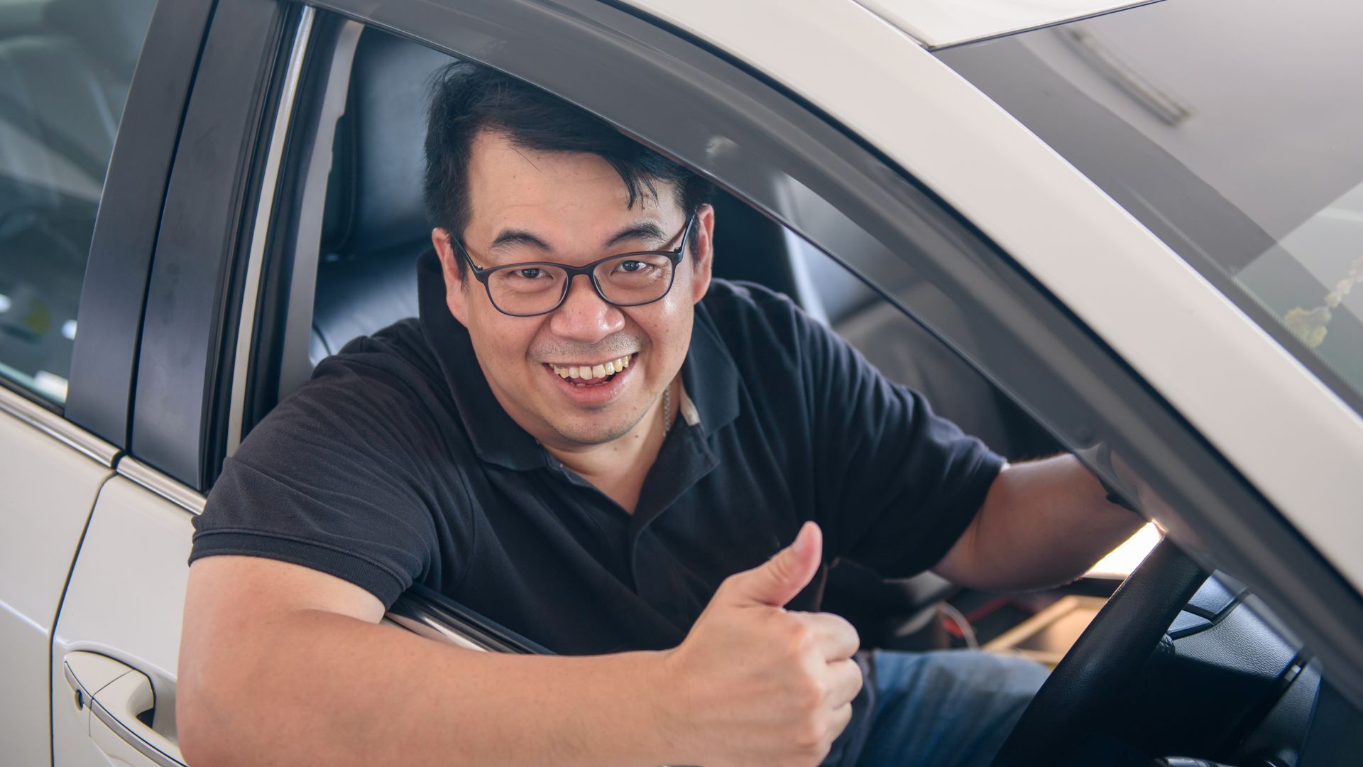 Smiling driver in a black shirt giving a thumbs-up inside a car