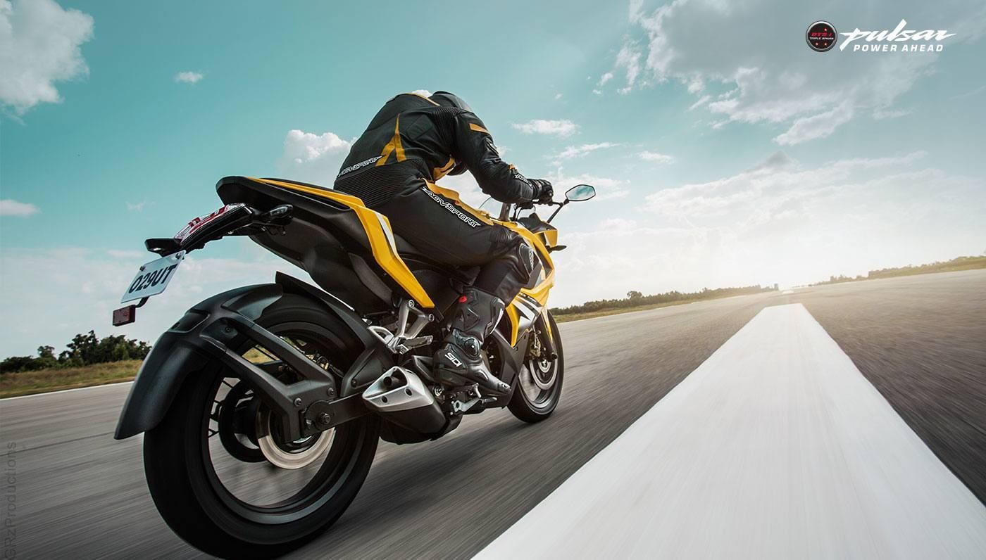 Yellow Bajaj motorcycle on road with rider, under a blue sky with clouds.