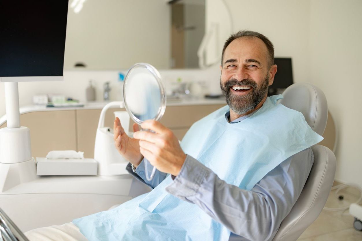 Man smiles while looking at a mirror in a dentist's chair. He wears a bib. Dental office setting.