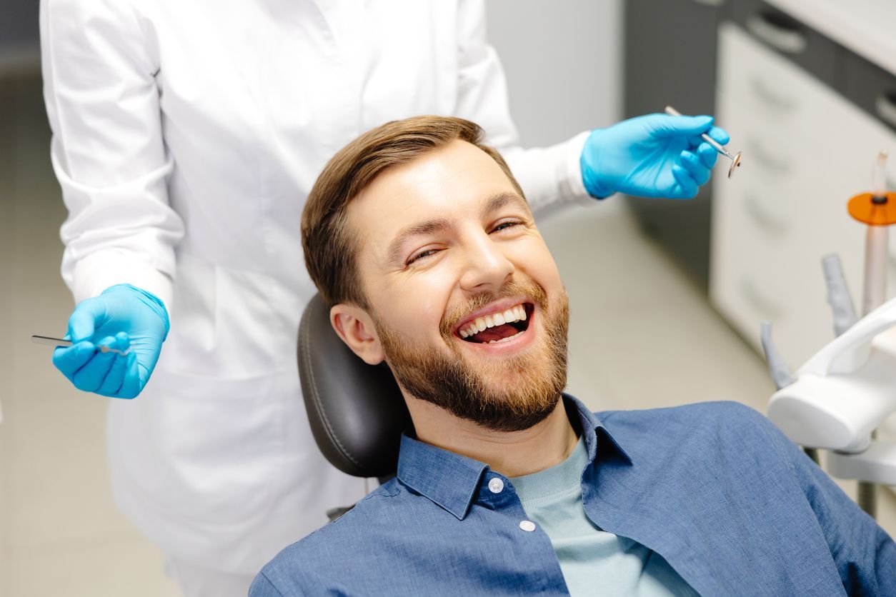 Man laughing in a dentist chair, dentist in gloves and coat holding tools.