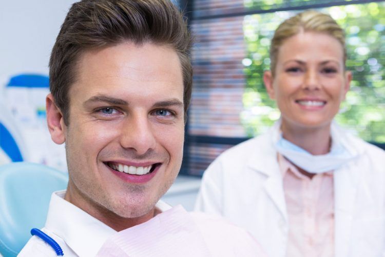 Smiling patient in dental chair, dentist in background. Dental office setting, bright and clean.