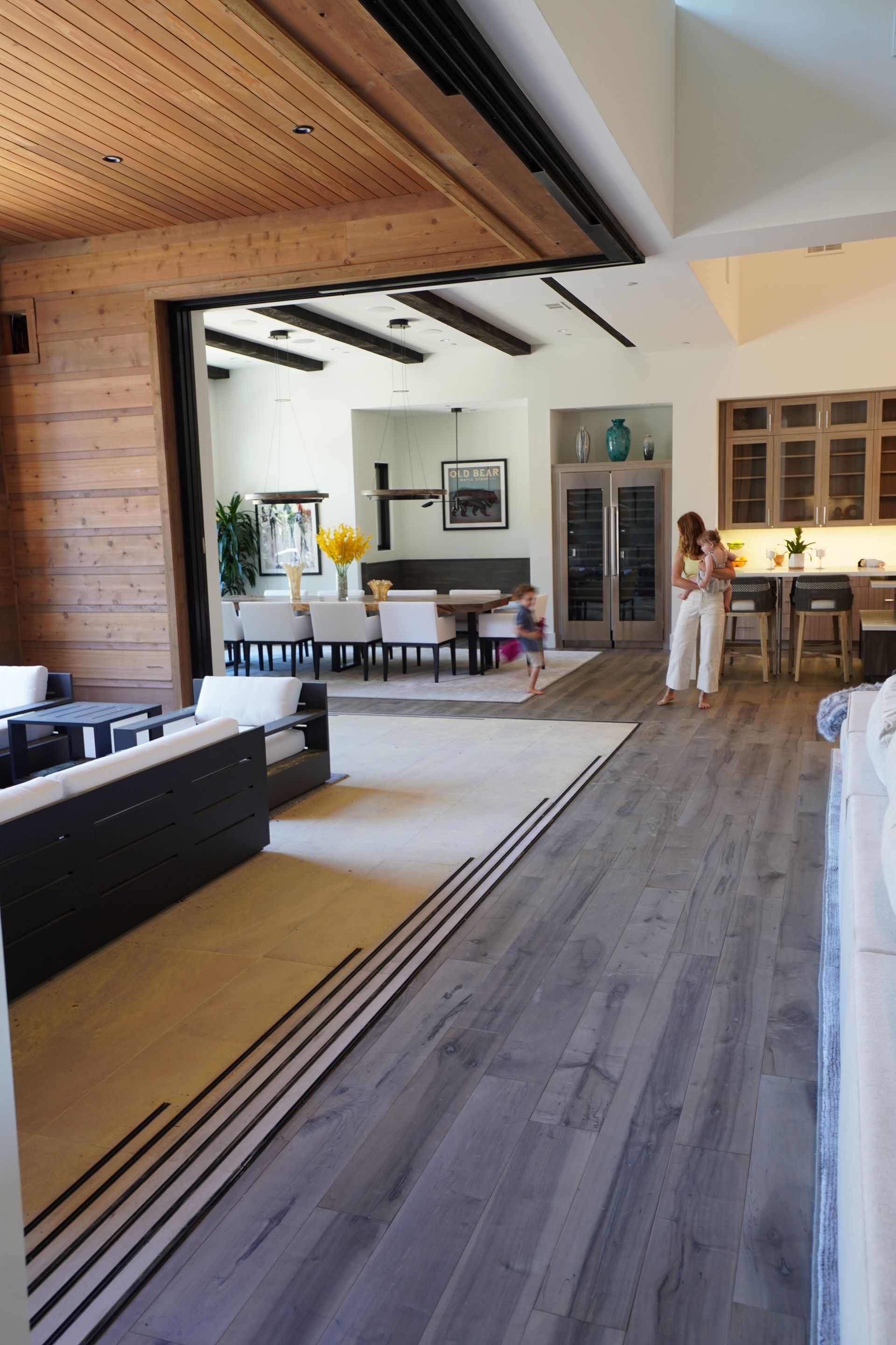 Woman holding baby and watching a child run through a large open door living room to the outdoors.