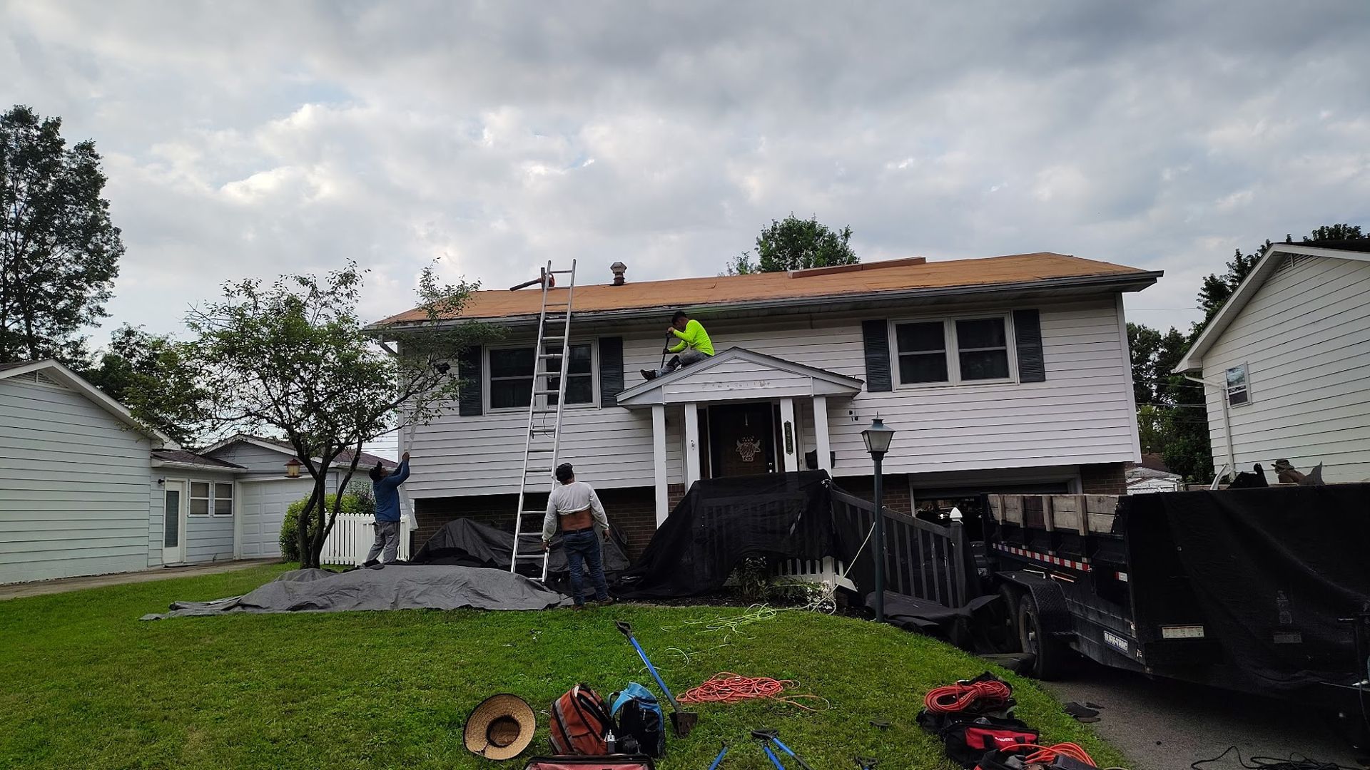 Workers on roof of white house, replacing shingles. Ladders and debris on lawn. Overcast day.