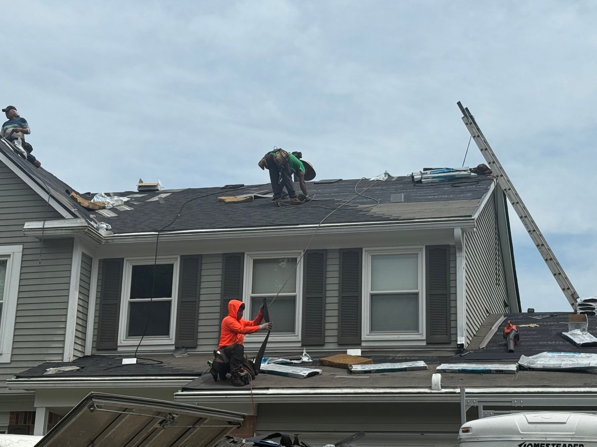 Roofers removing and replacing shingles on a two-story house with gray siding, under a cloudy sky.