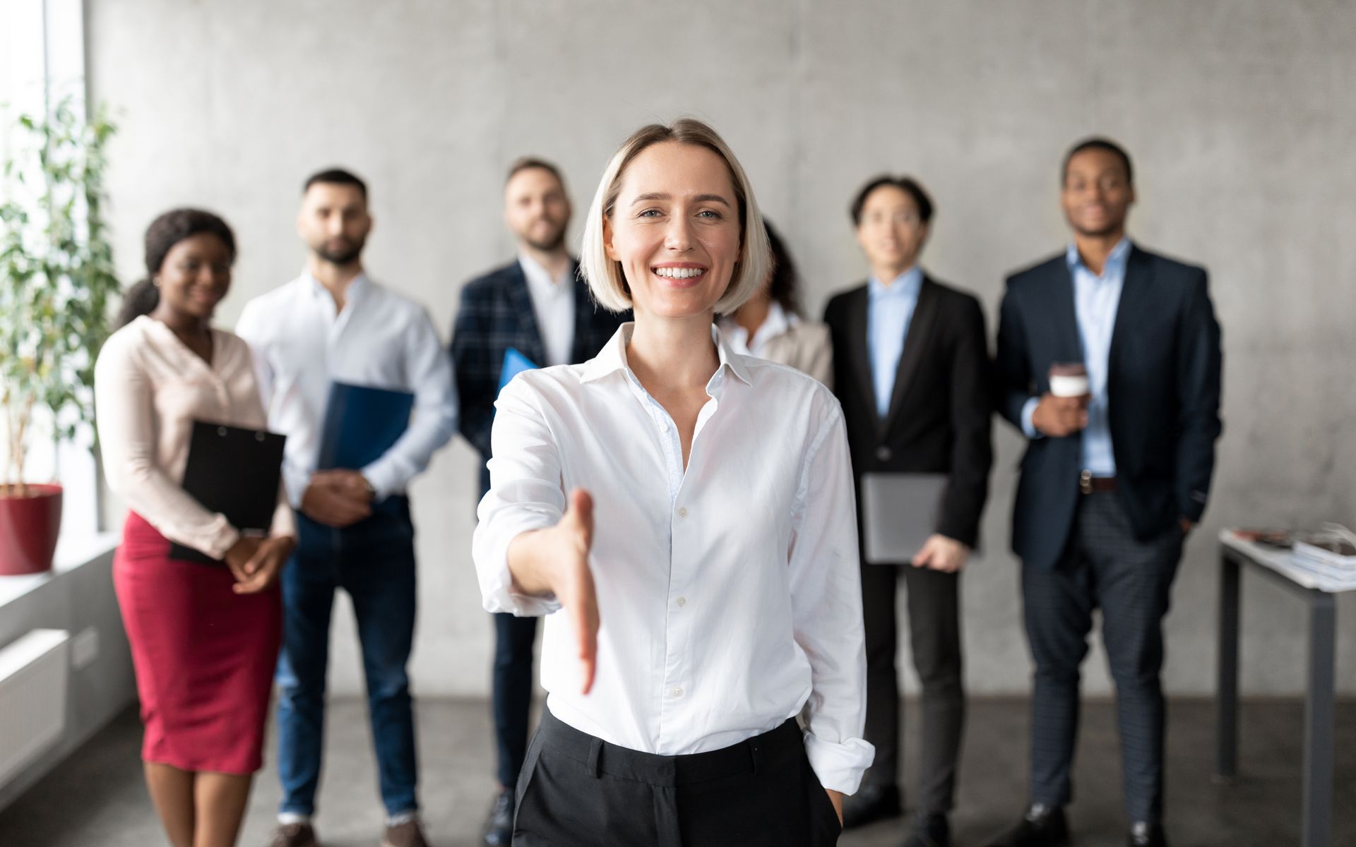 Smiling professional in white shirt shaking hands in office, with five colleagues standing behind her