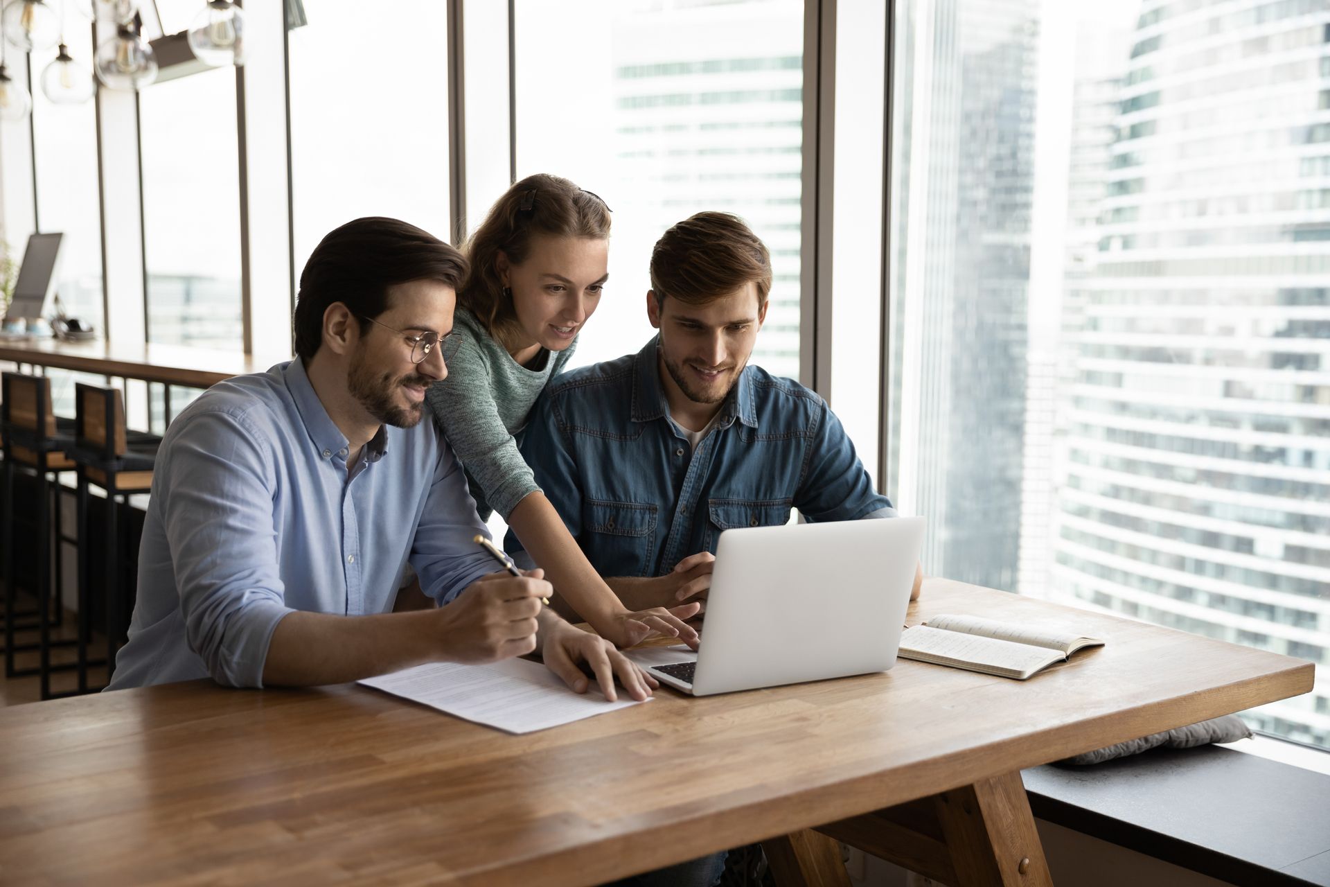 Three coworkers collaborating at a laptop in a bright office by large windows