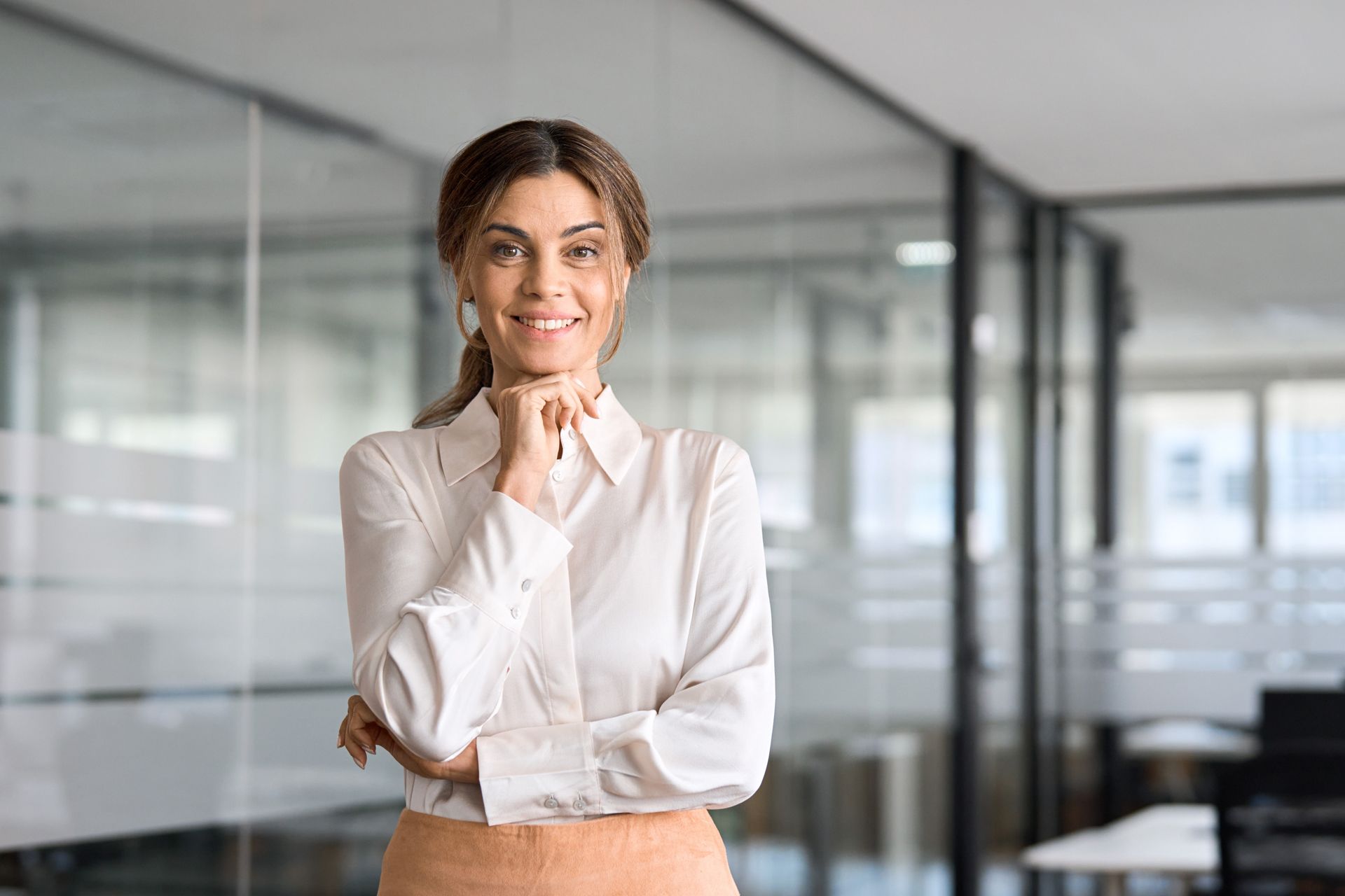 Woman in a white blouse stands in a modern office, smiling with her hand on her chin.