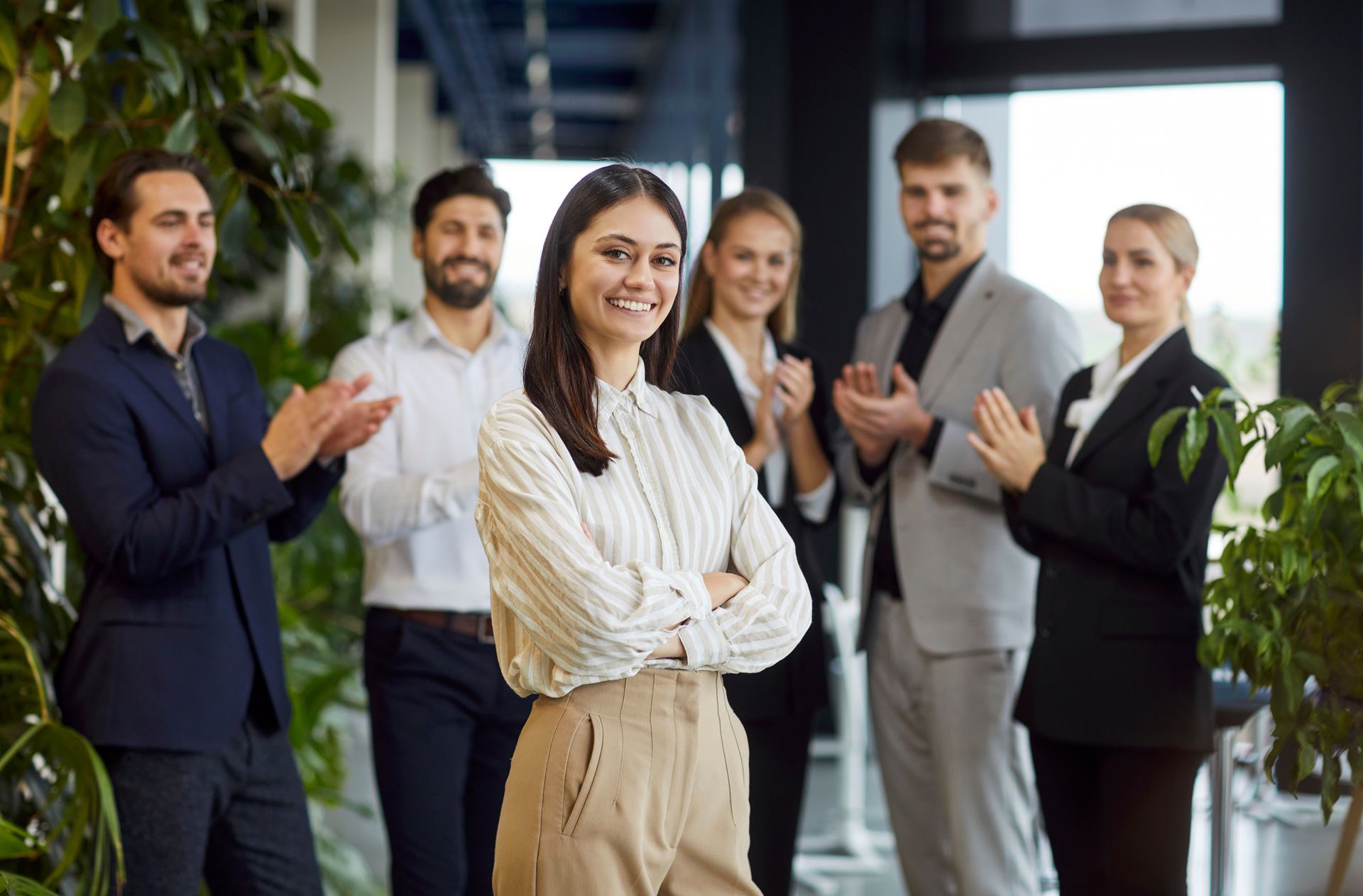 Team of professionals standing in an office, smiling and applauding, with a woman in a white blouse in front.