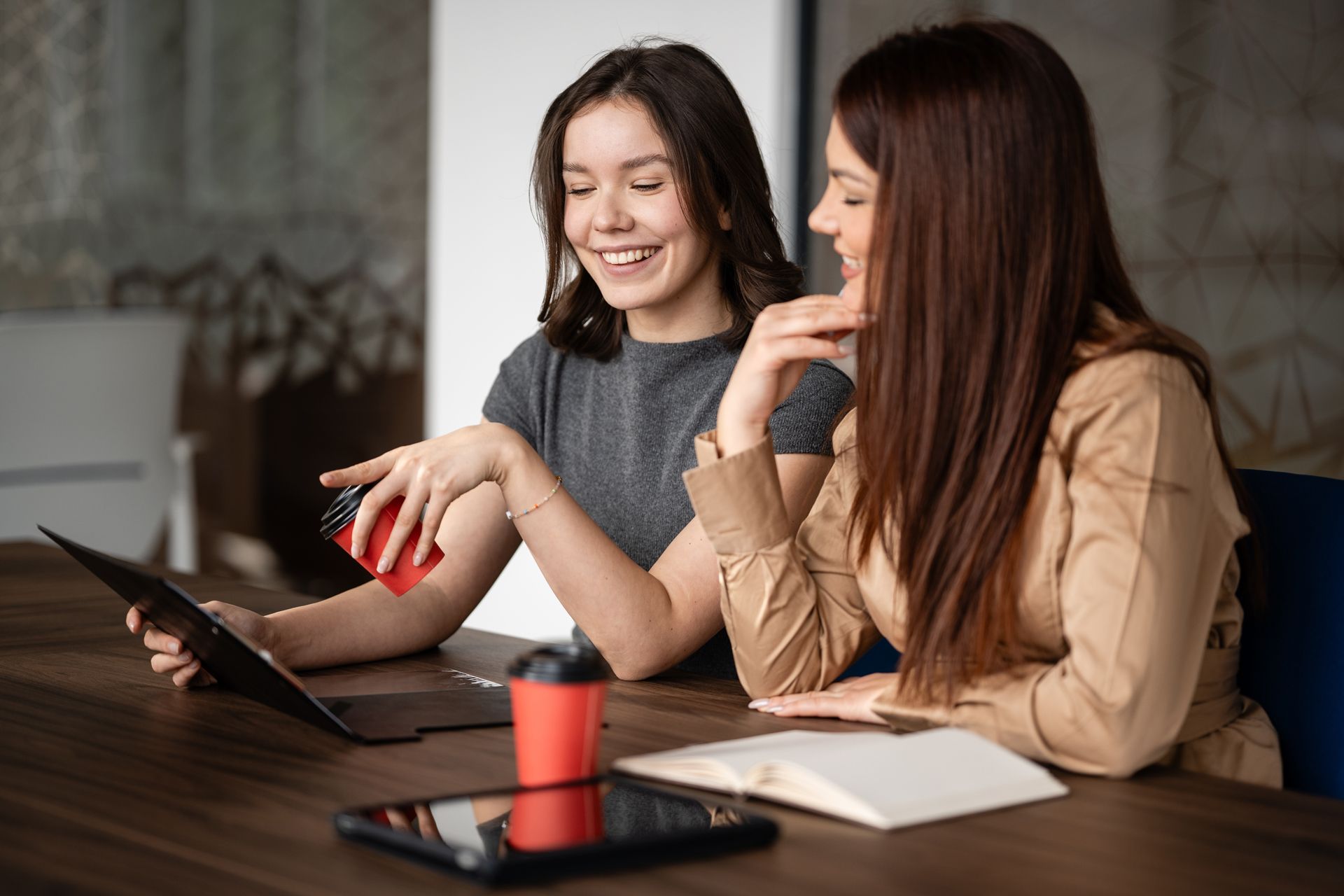 Two women at a table reviewing a laptop, with a notebook and red mug in the foreground.