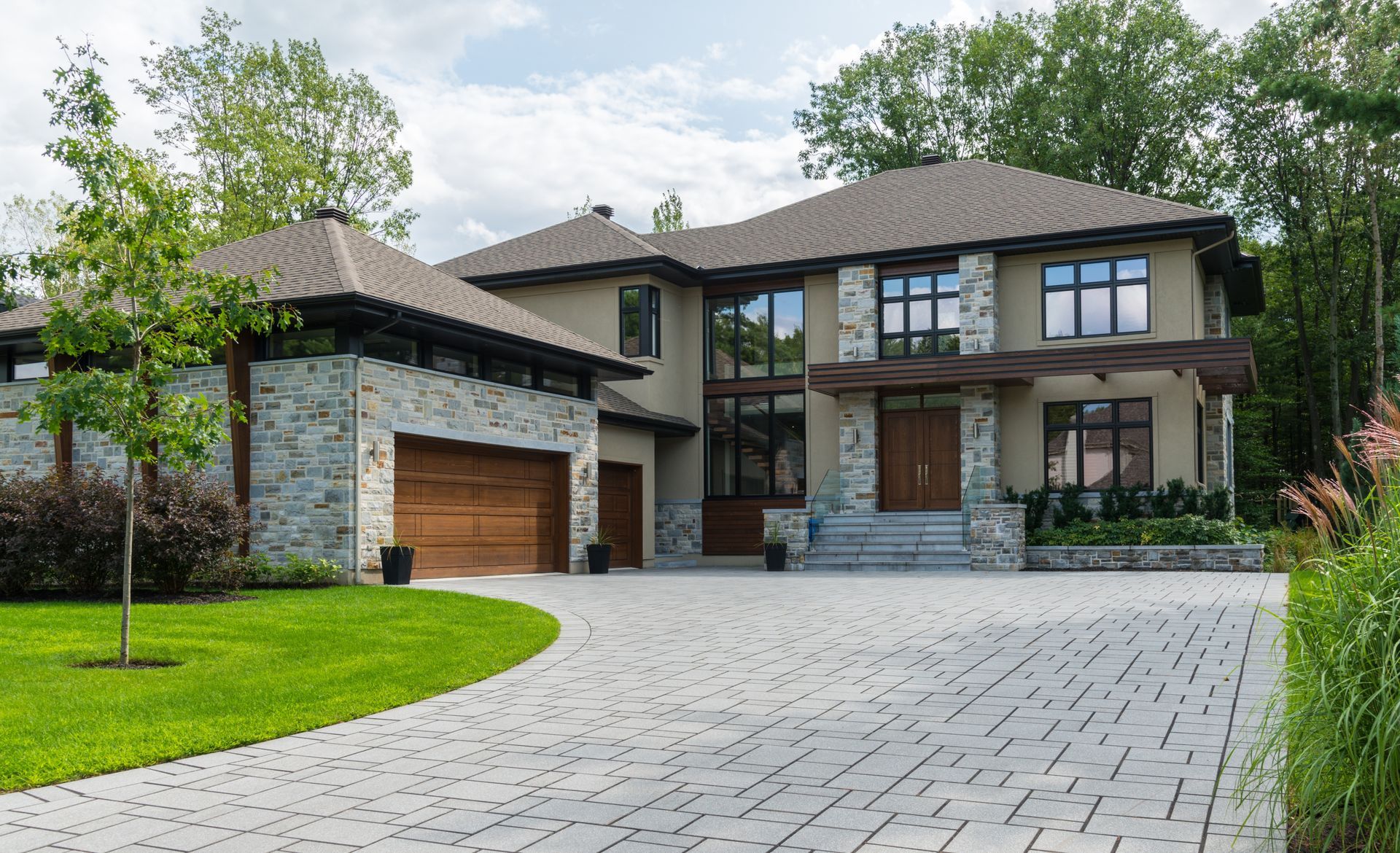 A two-story stone and stucco house with a wooden double garage, a paver driveway, and a well-manicured green lawn.