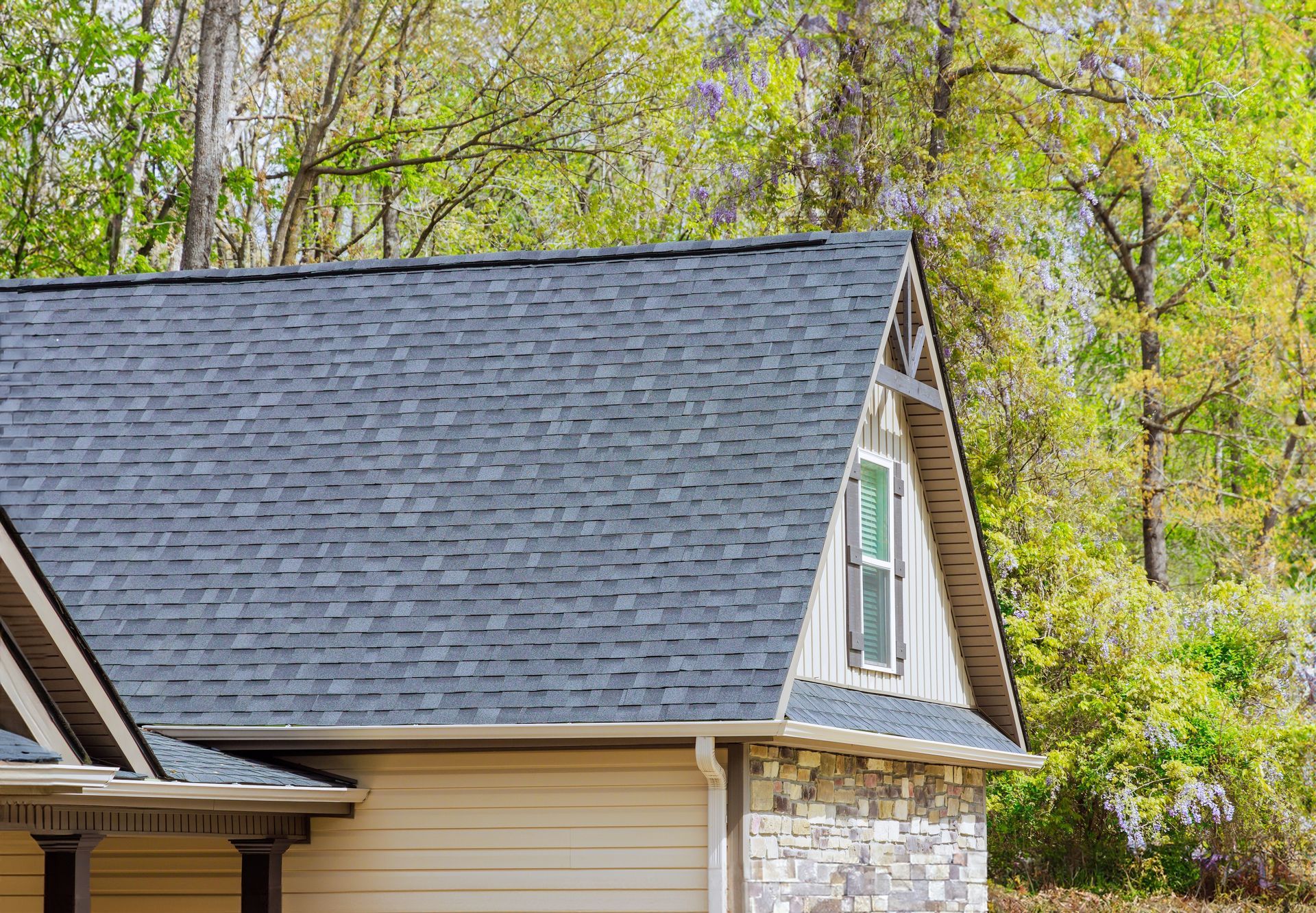 House roof with dark gray shingles, beige siding, and a dormer window against leafy trees
