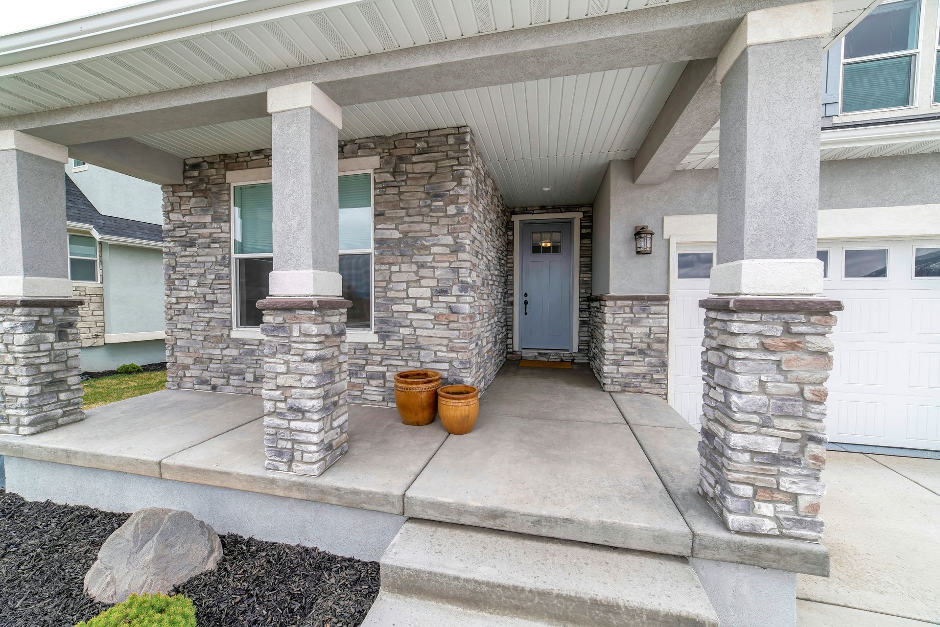 A front porch of a house with stone pillars, a light blue front door, two small orange pots, and a concrete entrance.