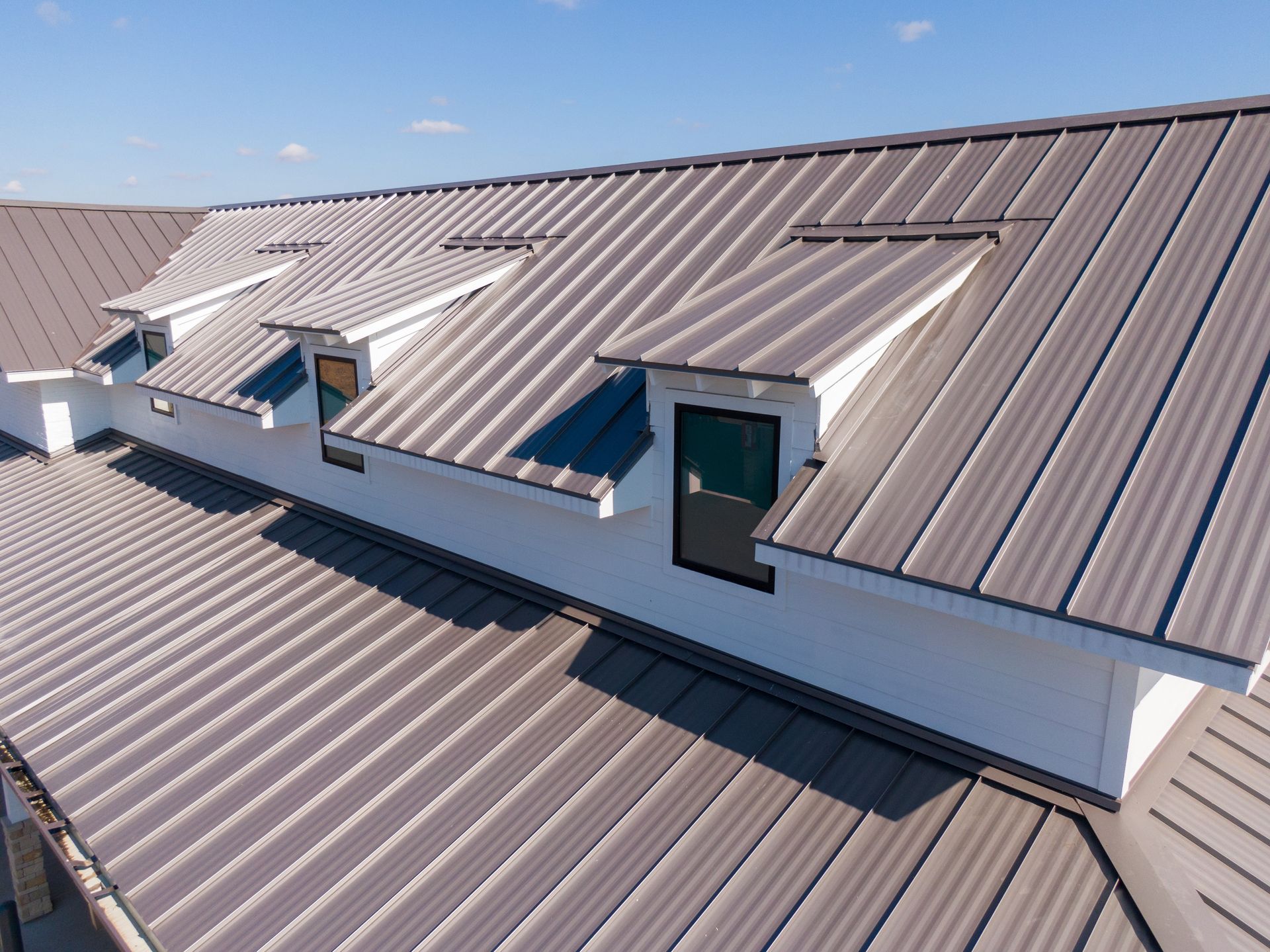 A brown standing-seam metal roof on a white building featuring three dormer windows under a clear blue sky.