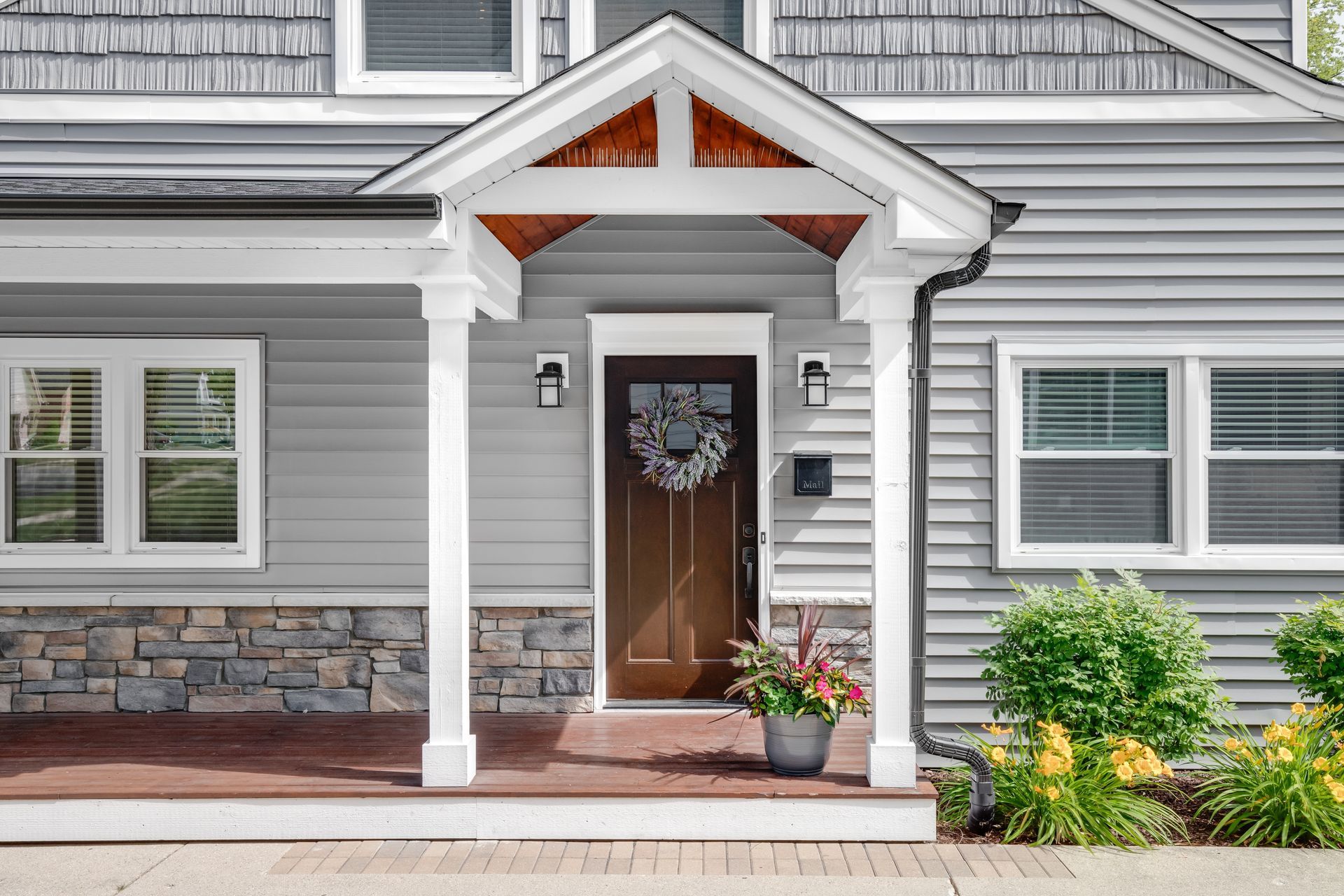 A suburban home entrance with gray siding, a brown front door with a wreath, a covered porch, and stone accents.