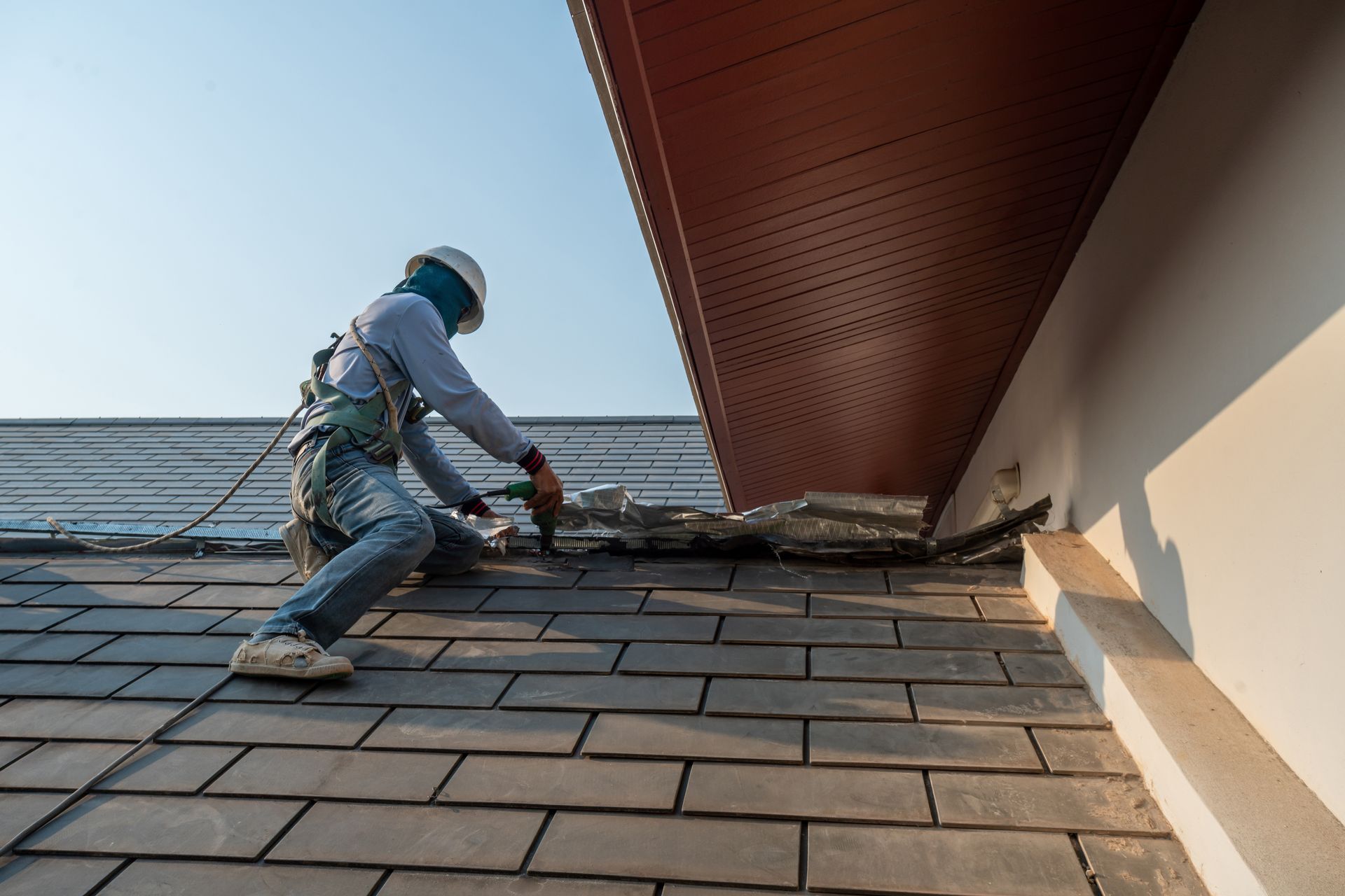 Roofer, wearing safety harness, repairing a damaged roof near a building's edge.