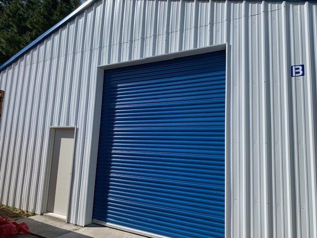 A metal warehouse exterior featuring a large blue roll-up door, a smaller adjacent white door, and the letter B.