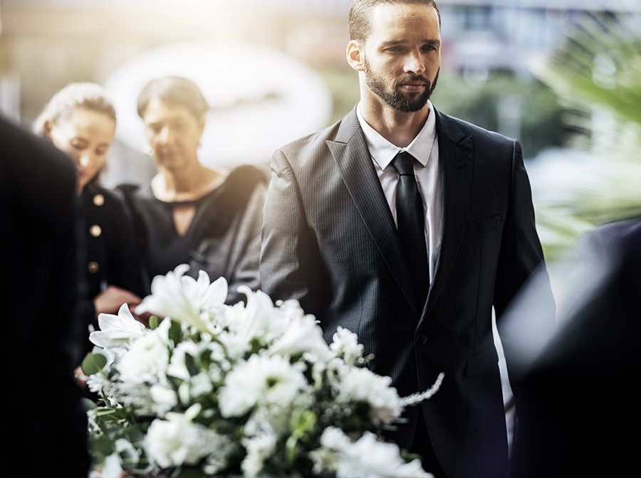 Person carrying casket with white flowers on top of casket and family in the back mourning loss of loved one