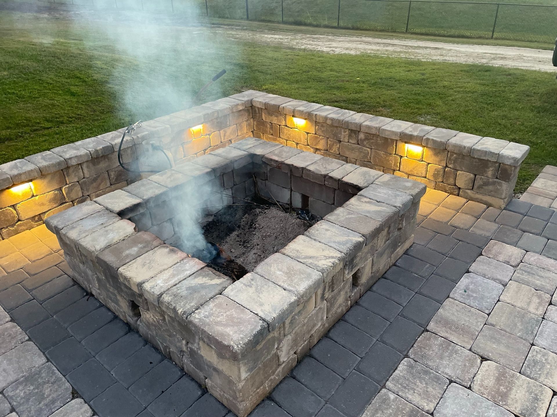 Stone fire pit with smoke rising, set in a paved backyard patio with warm lights on the walls