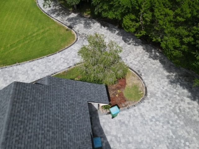 Aerial view of a gray roof, curved stone driveway, green lawn, and trees beside a patio chair.