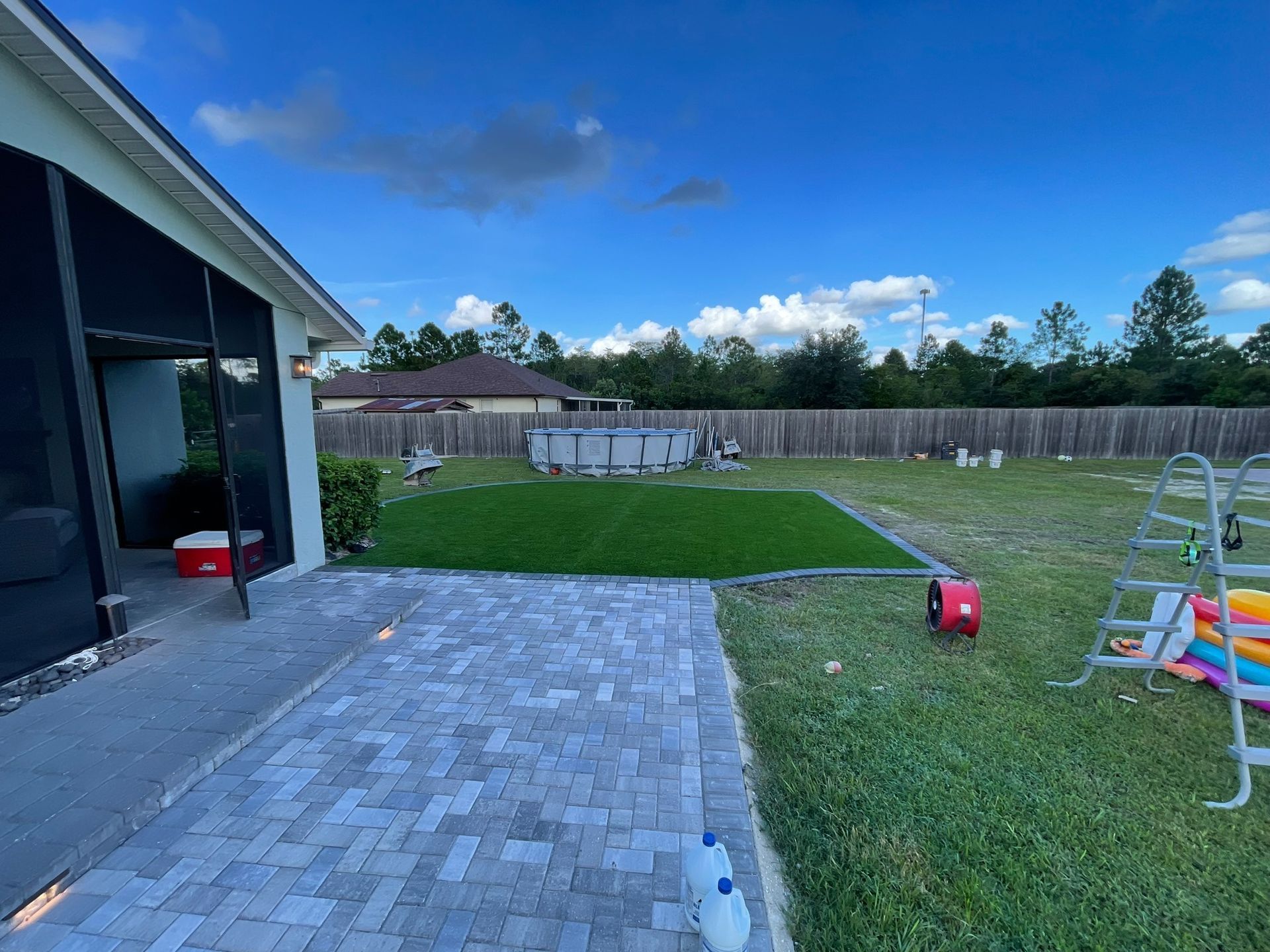 Backyard patio with a paved area, green lawn, pool, and children’s play equipment under a blue sky