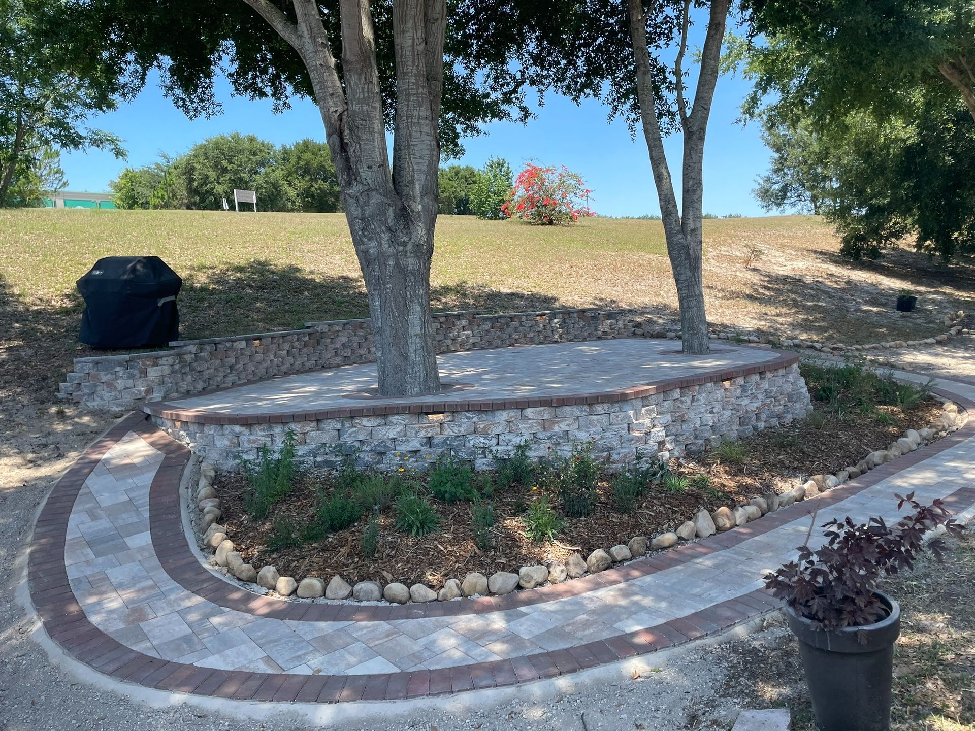 Curved stone garden bed with trees, rocks, and a small potted plant on a grassy hillside.