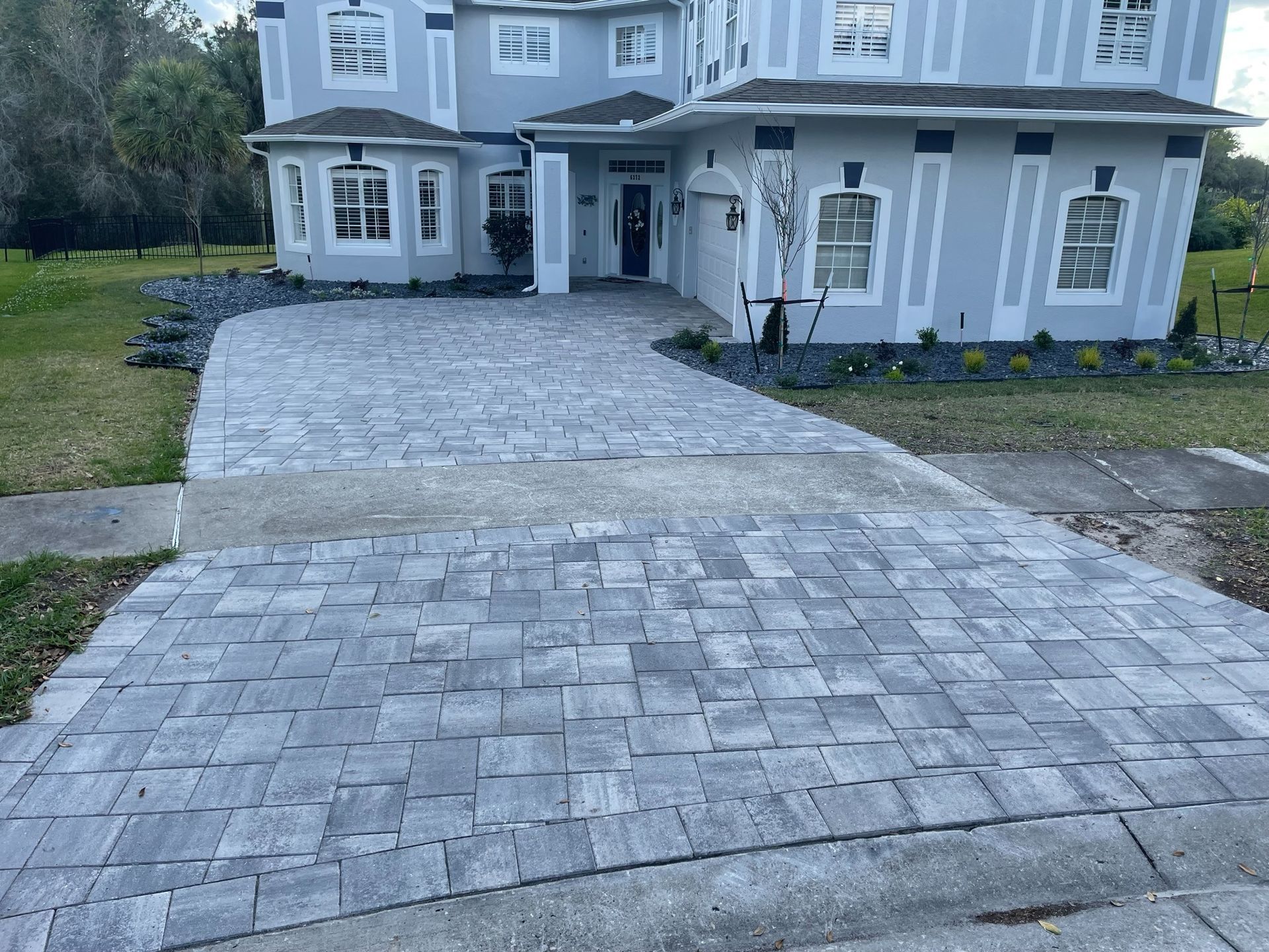 Stone-paver driveway leading to a large gray house with white trim and bay windows.