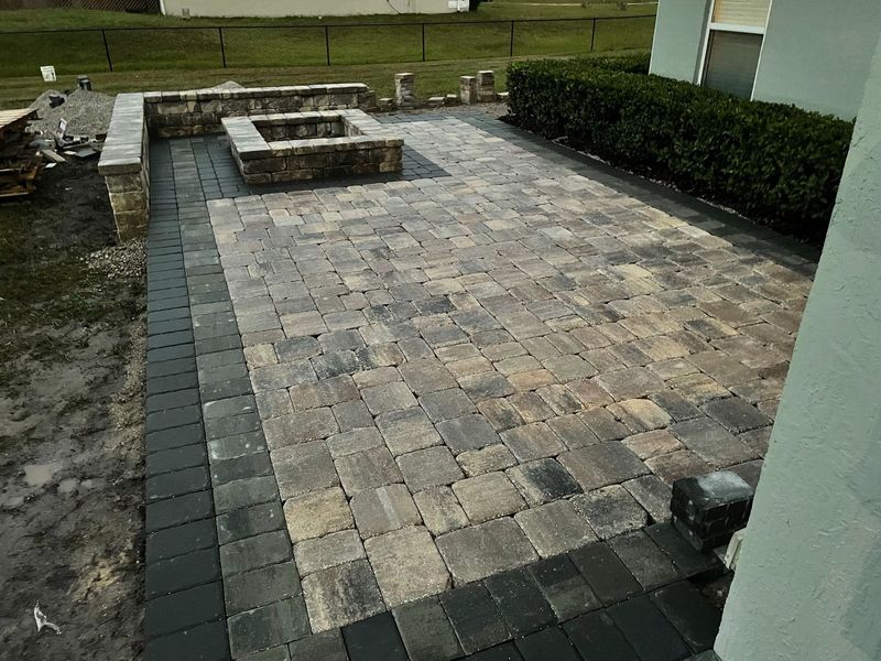Stone-paver patio with a raised fire pit, bordered by dark edging and shrubs beside a house.