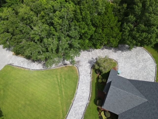 Aerial view of a driveway beside green lawn, dense trees, and a gray-roofed house.