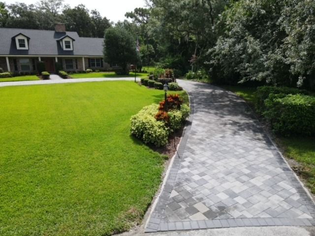 Curved gray paver driveway beside a green lawn and landscaped shrubs leading to a house