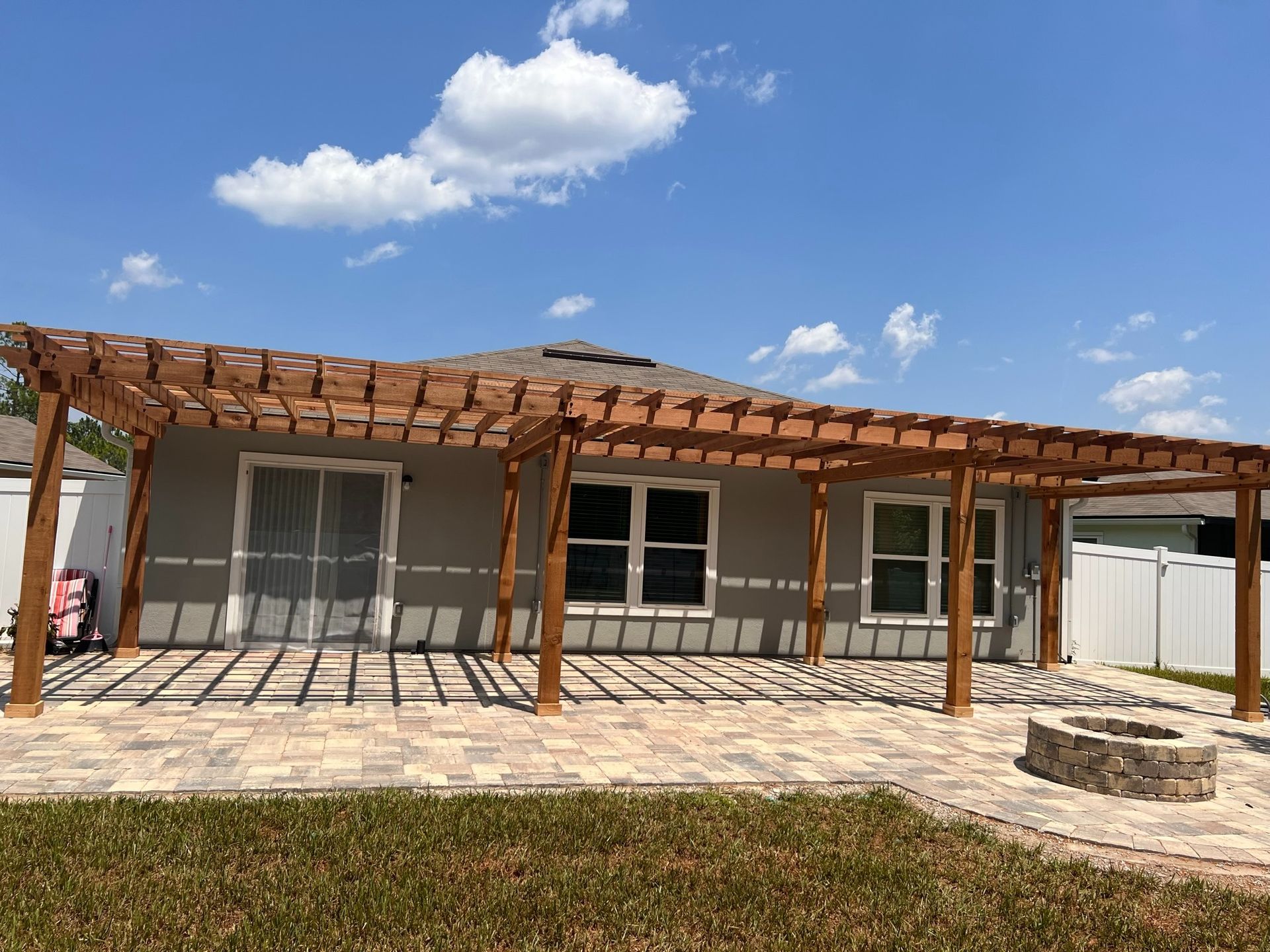 Backyard patio with a wooden pergola, sliding glass door, and small fire pit under a blue sky