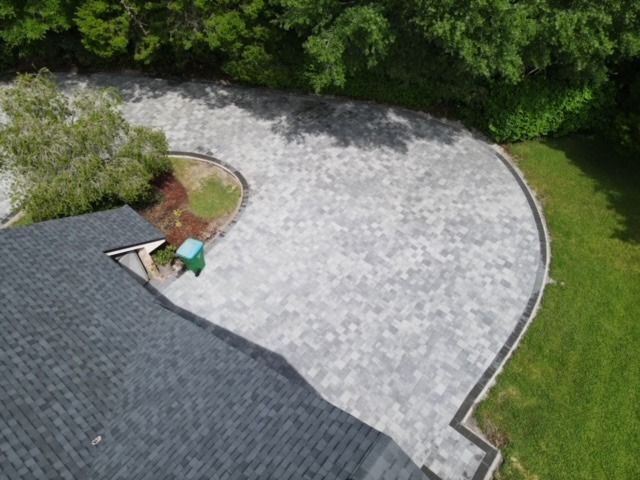 Aerial view of a curved gray driveway beside green lawn, trees, and a dark roof edge.