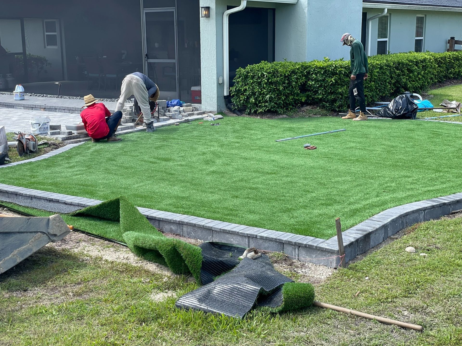 Workers replacing sod in a front yard beside a house, with rolled turf and tools on the grass.