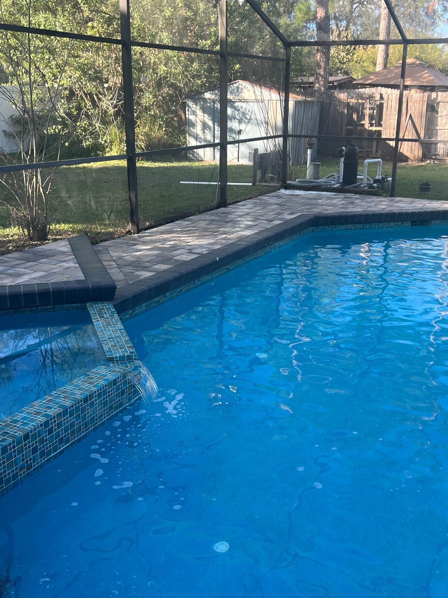 Backyard swimming pool with blue water, stone coping, and a fenced grassy yard in the background